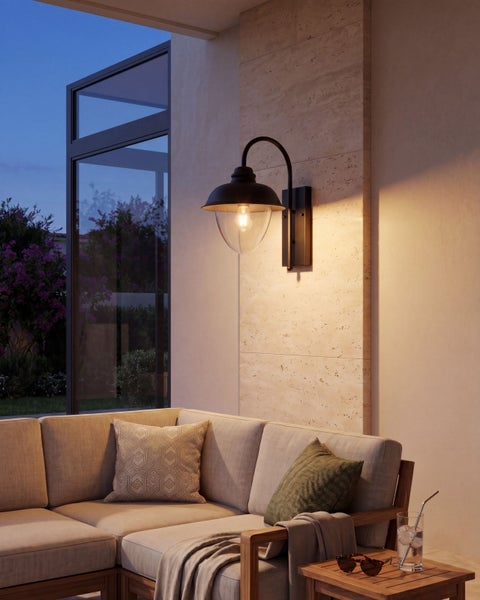 Außenwandleuchte mit gebogenem Arm und Glasschirm an einer Steinwand über einem Außensofa im Abendlicht.