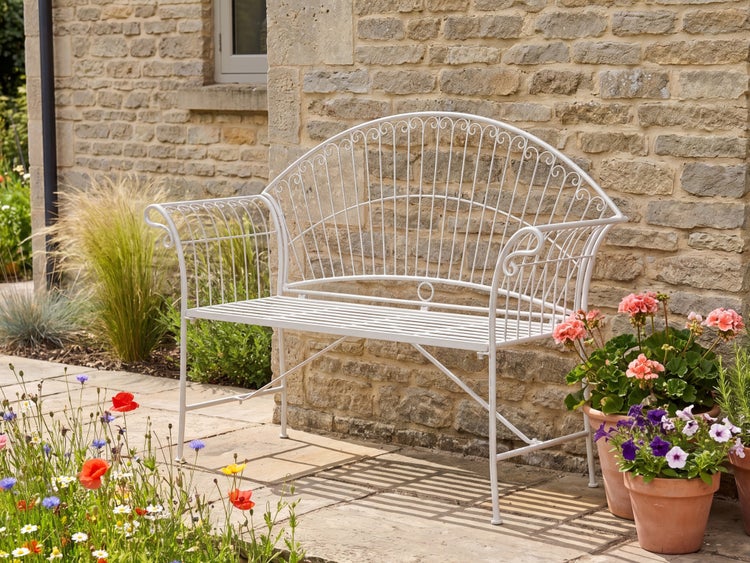 Filigrane Gartenbank aus Metall vor einer Natursteinmauer, umgeben von blühenden Pflanzen auf einer Terrasse.