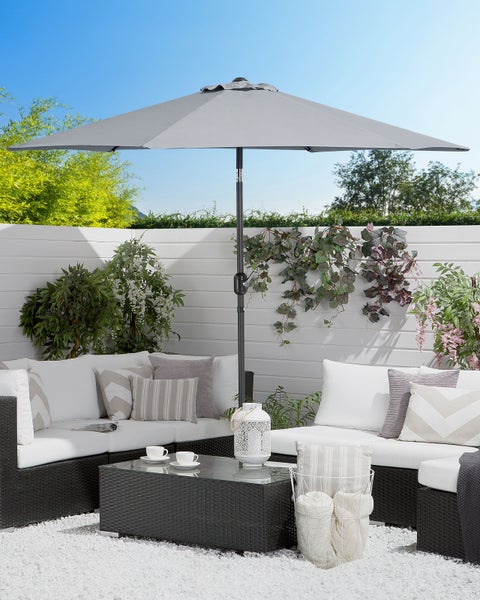 Grauer Sonnenschirm über einer hellen Gartenlounge mit dunklen Polyrattan-Möbeln, weißen Kissen und einem Couchtisch auf Kies.