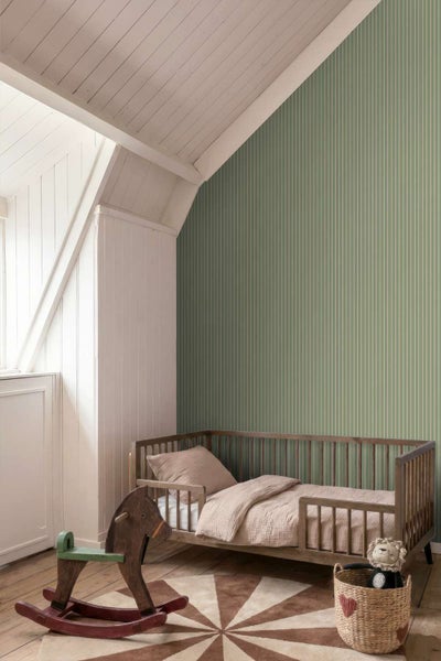 Ein Kinderzimmer mit einer Vliestapete mit vertikalen Streifen, einem Holzgitterbett und einem hölzernen Schaukelpferd auf einem runden Teppich.
