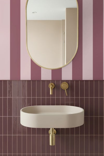 Modernes Badezimmer mit ovalem beigefarbenem Waschbecken, goldener Armatur und ovalem Spiegel vor gestreifter Tapete und dunklen Fliesen.