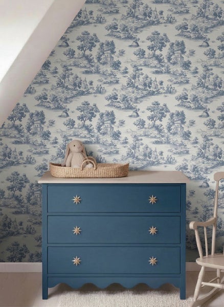 Vlies-Tapete mit klassischem Toile-de-Jouy-Muster in einem hellen Kinderzimmer hinter einer Kommode mit Weidenkorb und Stoffhase.