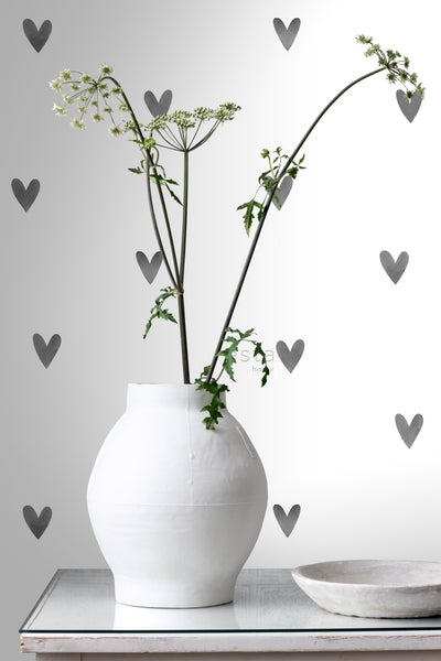 Dekorative Szene mit einer weißen Vase und Pflanzen auf einem Tisch vor einer Wand mit Herzmotiven.