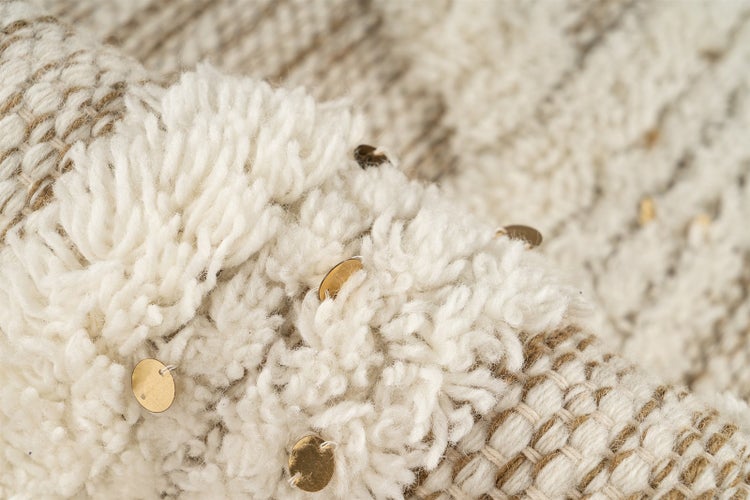 Nahaufnahme eines Teppichs mit zotteligem Flor und kleinen, runden Pailletten in Goldoptik auf einem flachgewebten Untergrund.