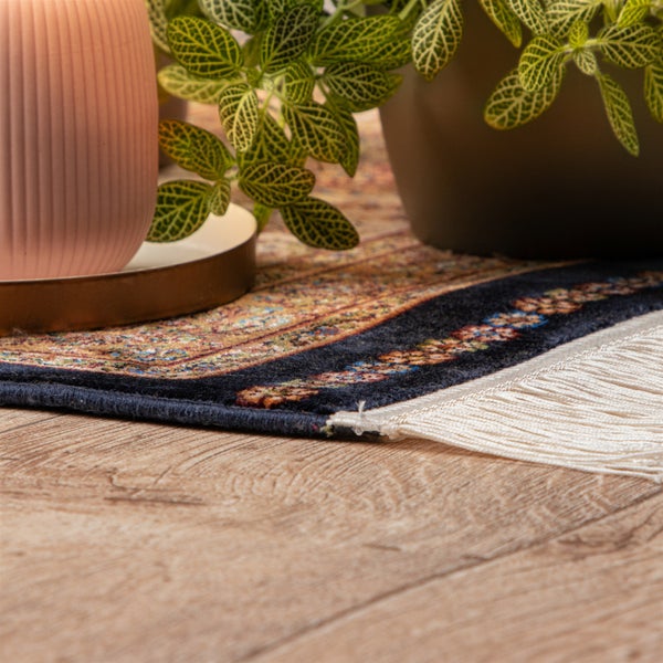 Nahaufnahme eines gemusterten Teppichs mit weißen Fransen auf hellem Holzboden, im Hintergrund eine geriffelte Vase und eine Pflanze.