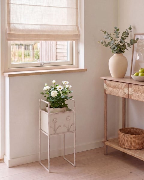 Helle Inneneinrichtung mit Fenster, Raffrollo, einem weißen Metall-Blumenständer und einem Konsolentisch aus Holz und Geflecht.