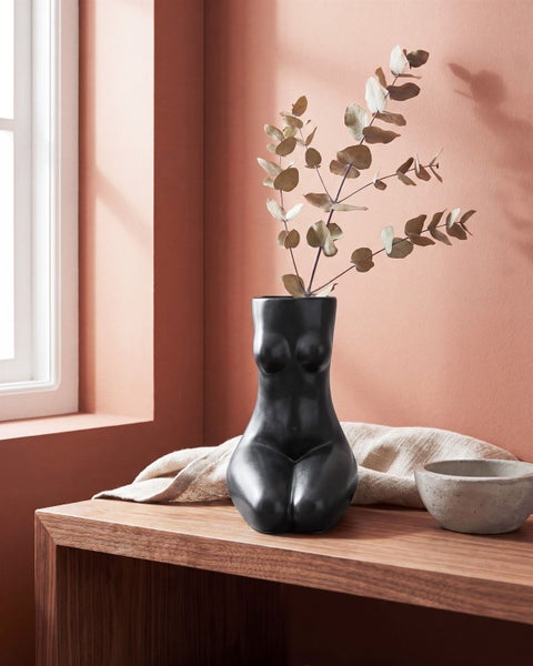 Dekorative Vase in Frauenkörper Form auf einem Holzregal mit Trockenblumen und einer Betonschale als Wohnaccessoire.