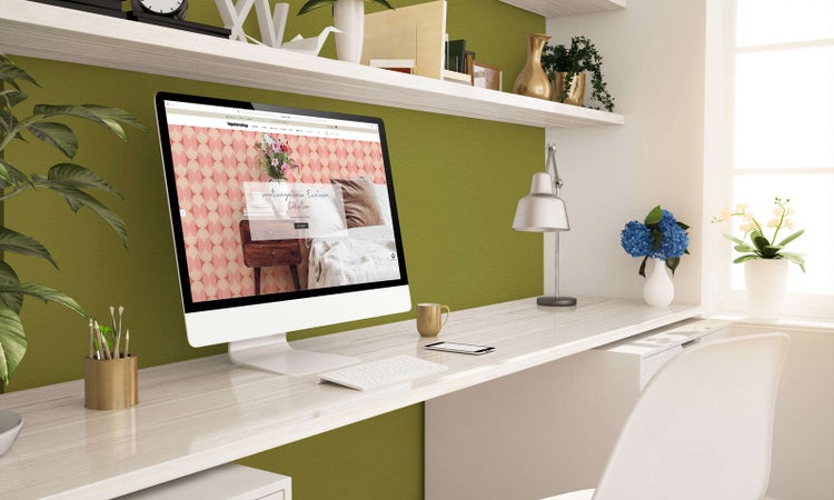 Hell eingerichteter Arbeitsplatz mit weißem Schreibtisch, Monitor, olivgrüner Wand und Wandregalen aus Holz.