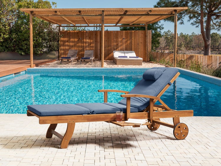 Gartenliege aus Holz mit blauer Auflage und Rädern auf einer Terrasse am Pool, im Hintergrund eine Holzpergola.