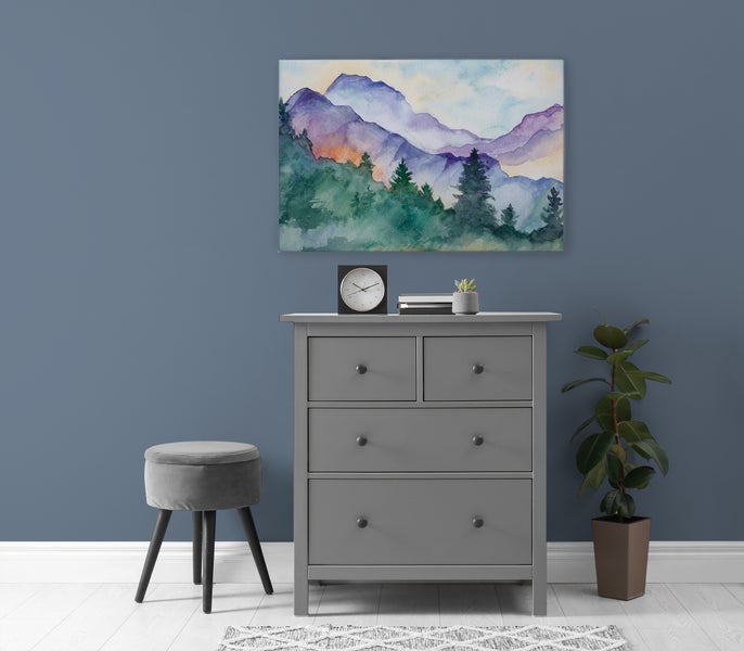 Wohnraum mit grauer Kommode, gepolstertem Hocker, Zimmerpflanze und einem Wandbild mit einer Berglandschaft im Aquarellstil.