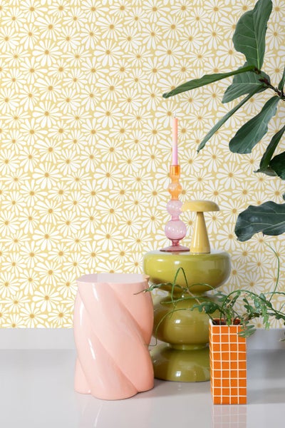 Vliestapete mit weißem Gänseblümchen-Muster auf gelbem Grund in einem hellen Raum mit farbenfrohen Design-Objekten und Zimmerpflanzen.