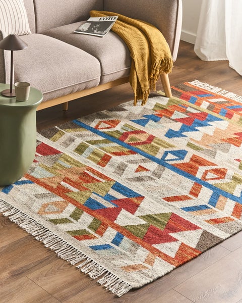 Bunter Teppich mit geometrischem Muster und Fransen vor einem beigen Sofa in einem hellen Wohnzimmer mit Holzboden.