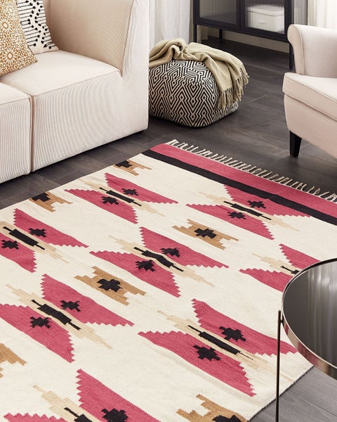 Moderner Kelim-Teppich mit pinkfarbenen, schwarzen und beigen geometrischen Mustern und Fransen, auf dunklem Boden in einem hellen Wohnzimmer.