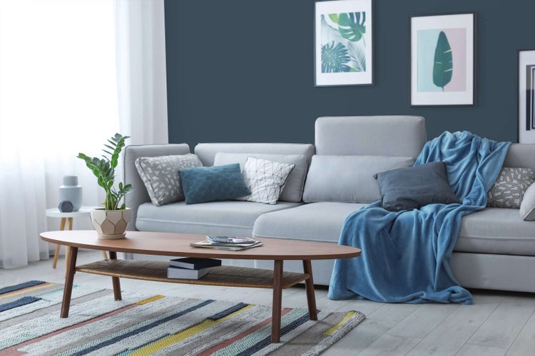Modernes Wohnzimmer mit dunkelblauer Wandfarbe, hellgrauem Ecksofa, ovalem Couchtisch aus Holz und einem gestreiften Teppich.