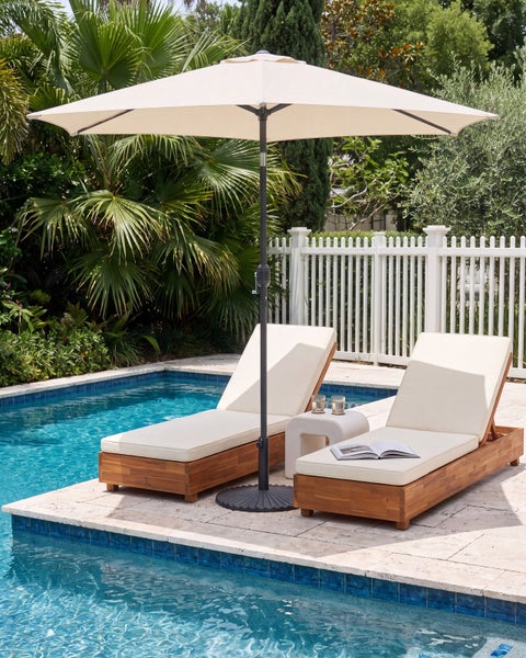 Zwei Gartenliegen aus Holz mit Polstern und ein großer Sonnenschirm an einem Pool im Garten mit Palmen und einem weißen Zaun.
