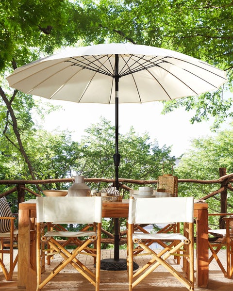 Großer cremefarbener Sonnenschirm über einem hölzernen Esstisch mit Regiestühlen auf einer hellen, von Bäumen umgebenen Terrasse.