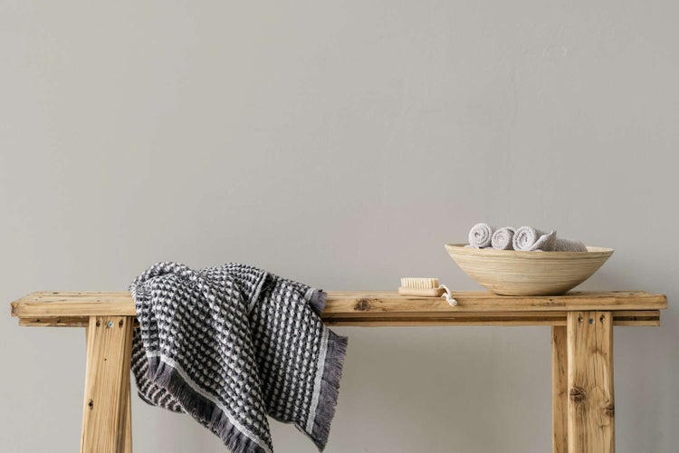 Strukturiertes Handtuch mit Fransen und Waffelmuster auf einer rustikalen Holzbank neben einer Schüssel mit Handtüchern und einer Bürste.