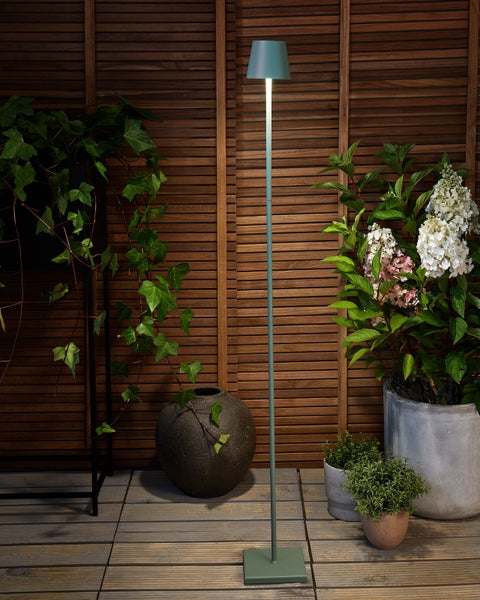 Schlanke grüne Stehleuchte für den Außenbereich auf einer Holzterrasse vor einer Lamellenwand umgeben von Pflanzen wie Efeu und Hortensien.