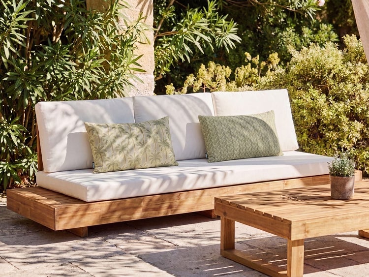 Gartensofa aus Holz mit hellen Sitzpolstern und zwei gemusterten Dekokissen in einer sonnigen Gartenumgebung.