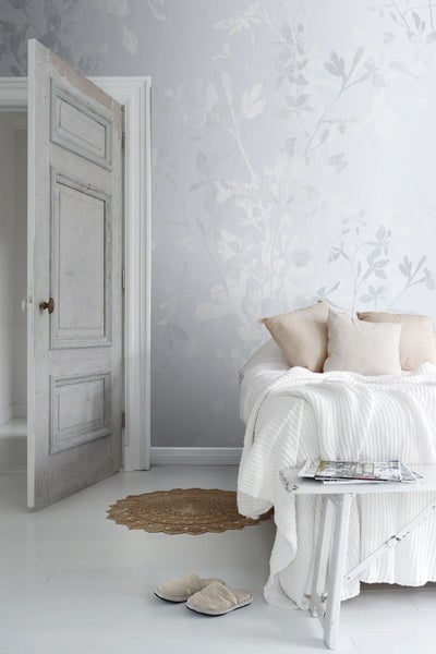 Helles Schlafzimmer mit floraler Tapete, geöffneter Holztür, Bett mit Kissen und Tagesdecke, Beistelltisch, Teppich und Hausschuhen.