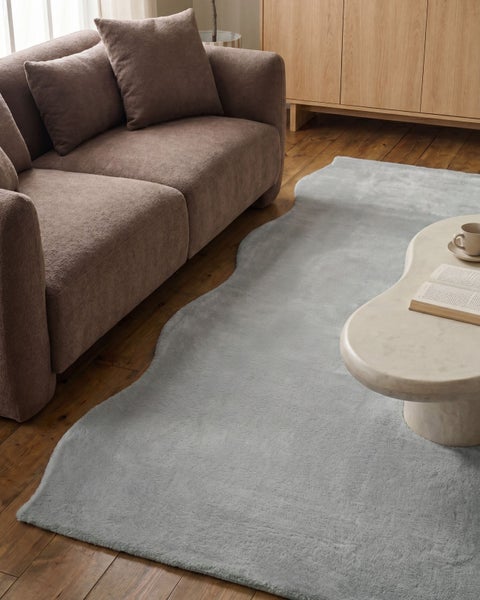 Weicher Teppich mit unregelmäßiger Form in einem Wohnzimmer auf Holzfußboden neben einem Sofa und einem Couchtisch aus Stein.