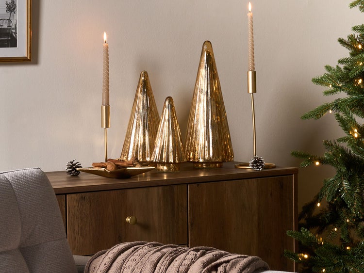 Dekoratives Arrangement mit goldenen Deko-Tannenbäumen, Kerzen in goldenen Kerzenständern und einem kleinen Dekotablett auf einer Holzkommode neben einem Weihnachtsbaum.