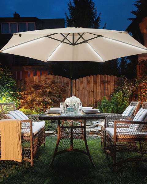 Großer beigefarbener Sonnenschirm mit LED-Beleuchtung über einer Sitzgruppe aus Polyrattan in einem beleuchteten Garten bei Nacht.
