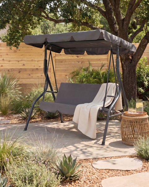 Hollywoodschaukel aus Metall mit verstellbarem Sonnendach und Sitzkissen auf einer Terrasse im Garten neben einem Beistelltisch.
