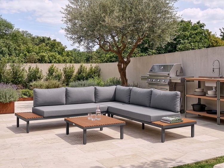 Moderne Lounge-Gartengruppe mit Ecksofa, Couchtisch und Ablageflächen aus Holz auf einer Steinterrasse mit Gasgrill und Außenküche im Garten.