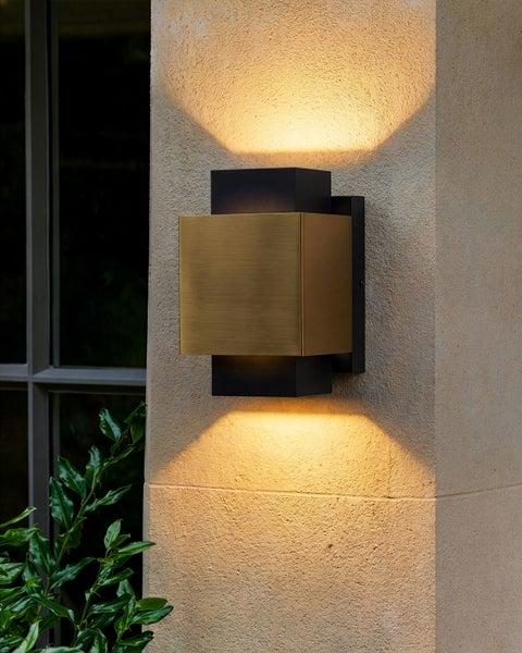 Rechteckige Außenwandleuchte aus Metall in Schwarz und Gold mit Licht nach oben und unten, montiert an einer hellen Steinwand neben einer Pflanze.