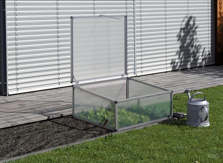 Ein offenes Frühbeet mit Aluminiumrahmen und Hohlkammerplatten auf einem Gartenbeet neben einer Rasenfläche und einer Gießkanne.