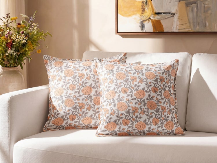 Zwei quadratische Zierkissen mit floralem Muster auf einem weißen Sofa neben einer Blumenvase.
