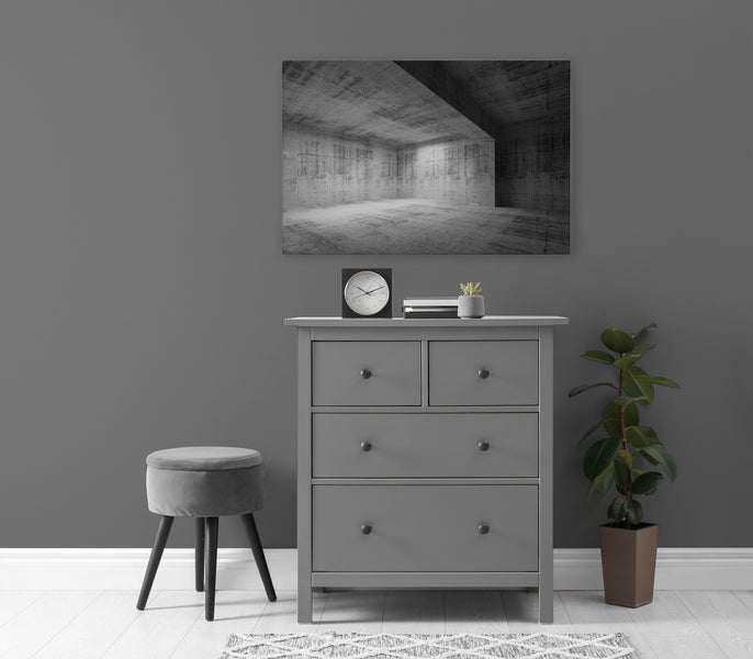 Eine Kommode mit vier Schubladen steht vor einer Wand mit einem Wandbild eines Betonraumes, daneben ein Hocker und eine Zimmerpflanze.