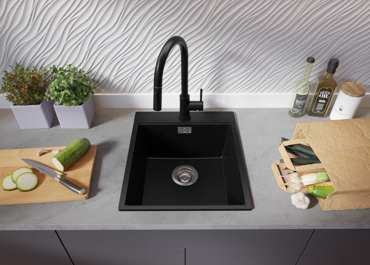 Schwarze Küchenspüle mit Wasserhahn, Gemüse, Kräutern und Olivenöl auf einer Arbeitsplatte.