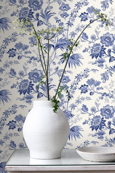 Florale Tapete mit blauem Blumen- und Blättermuster auf weißem Hintergrund, im Vordergrund eine weiße Vase mit grünen Zweigen.