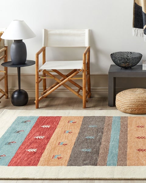 Bunter gestreifter Teppich mit Motiven in hellem Wohnzimmer mit Bambusstuhl, Lampe und Jute-Pouf auf Holzfußboden.