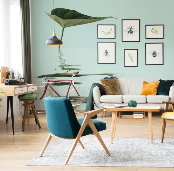 Hell eingerichtetes Wohnzimmer mit Fototapete mit Blattmotiv, Sofa, Couchtisch, Sessel, Schreibtisch und eingerahmten Wandbildern.