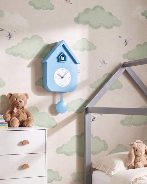 Wand-Kuckucksuhr in Hausform für Kinderzimmer vor einer Tapete mit Wolken und Vogelmotiv sowie Kuscheltieren und einem Hausbett.