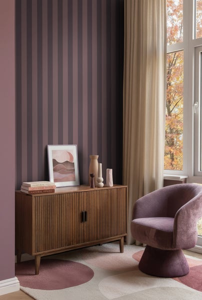 Modernes Wohnzimmer mit vertikal gestreifter Tapete, Sideboard aus Holz, Sessel und gemustertem Teppich vor einem Fenster.