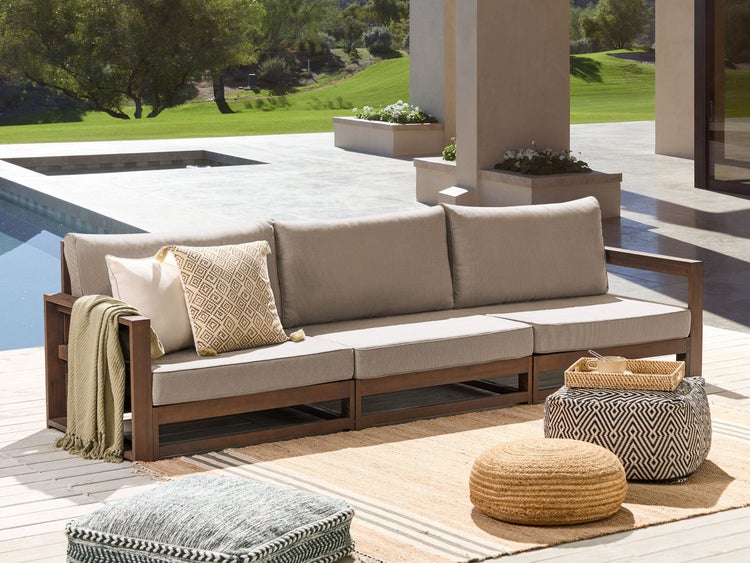 Im Bild ist eine Außenszene mit einem modularen Sofa, Poufs und einem Teppich zu sehen. Das Sofa hat einen Holzrahmen und Polster. Die Szene ist hell und einladend.