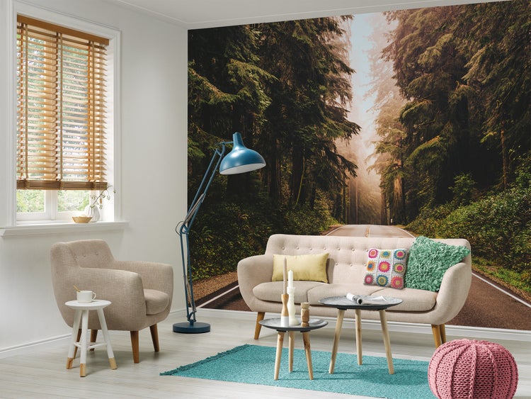 Helles Wohnzimmer mit großflächiger Fototapete einer Waldstraße, einem Sofa, einem Sessel, Couchtischen und einer blauen Stehlampe.