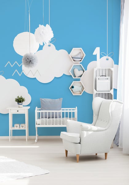 Hell gestaltetes Kinderzimmer mit Wolken-Wanddekoration, weißem Ohrensessel, Gitterbett und Beistelltisch auf hellem Boden.