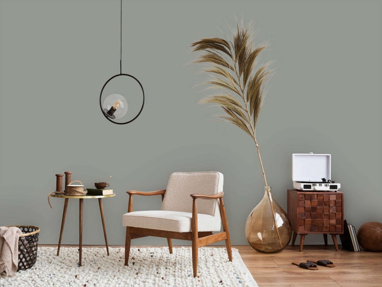 Helles Wohnzimmer mit salbeigrüner Wand, Polstersessel aus Holz, runder Metallbeistelltisch, Pendelleuchte und großer Glasvase mit Palmwedel.