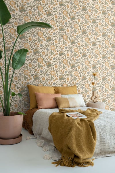 Schlafzimmer mit floral gemusterter Tapete, niedrigem Bett mit Kissen, Strickdecke und einer großen Zimmerpflanze.