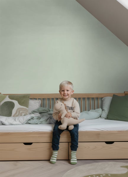 Helles Kinderzimmer mit hellgrün gestrichener Wand, Holzbett mit Schubladen und grünen Textilien.