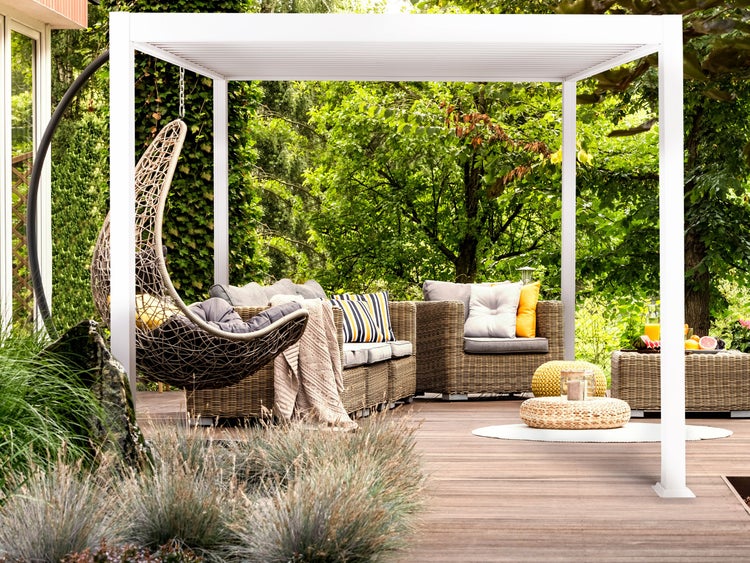 Weiße Pergola auf einer Holzterrasse mit Rattan-Lounge-Set, Hängesessel und Zierkissen in einer grünen Gartenumgebung.