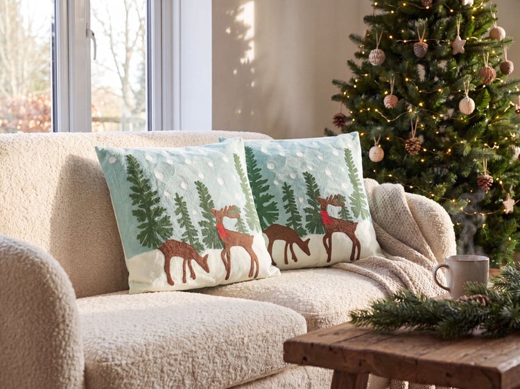 Zwei Dekokissen mit bestickten Winterszenen aus Rentieren und Bäumen auf einem strukturierten Sofa neben einem geschmückten Weihnachtsbaum.