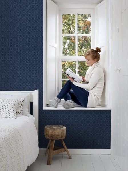 Wohnszene mit blau gemusterter Tapete, Bett, Fenster und lesender Person