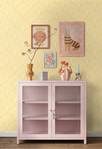 Rosa Schrank mit Glastüren vor einer gelben Mustertapete, dekoriert mit verschiedenen Vasen und Wandbildern im Retro-Stil.