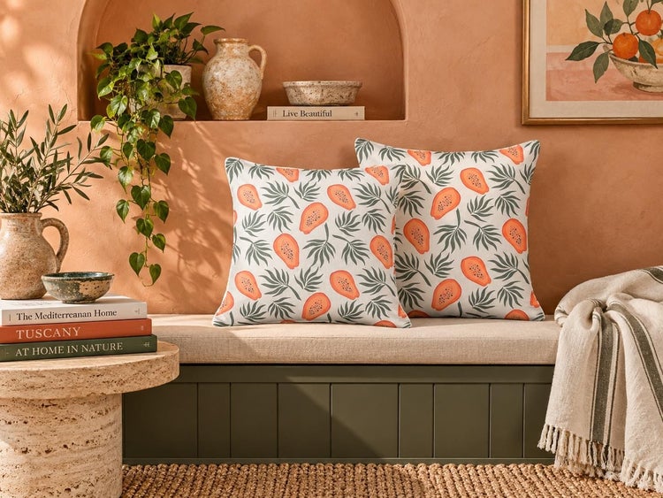 Zwei quadratische Dekokissen mit Papaya Motiv auf einer Sitzbank in einem mediterran eingerichteten Raum mit Pflanzen und Wohnaccessoires.